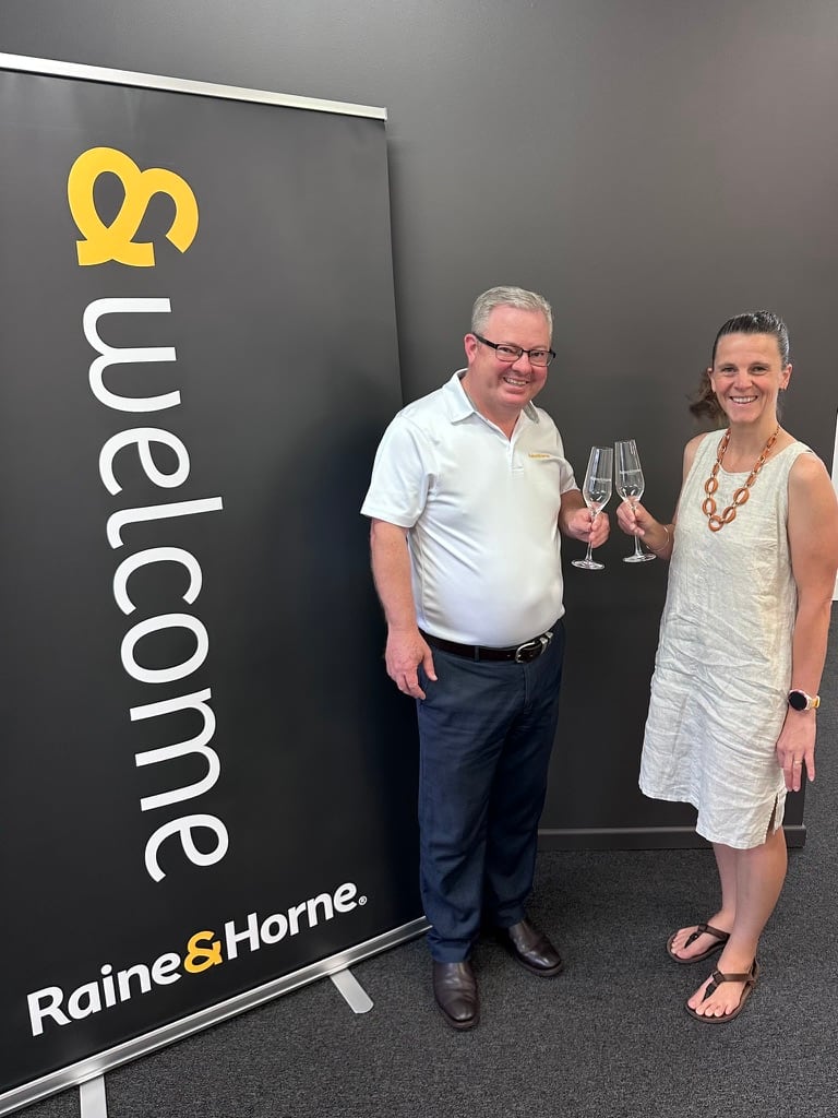 Chrissie and a Raine & Horne team member smiling and toasting with engraved champagne glasses
